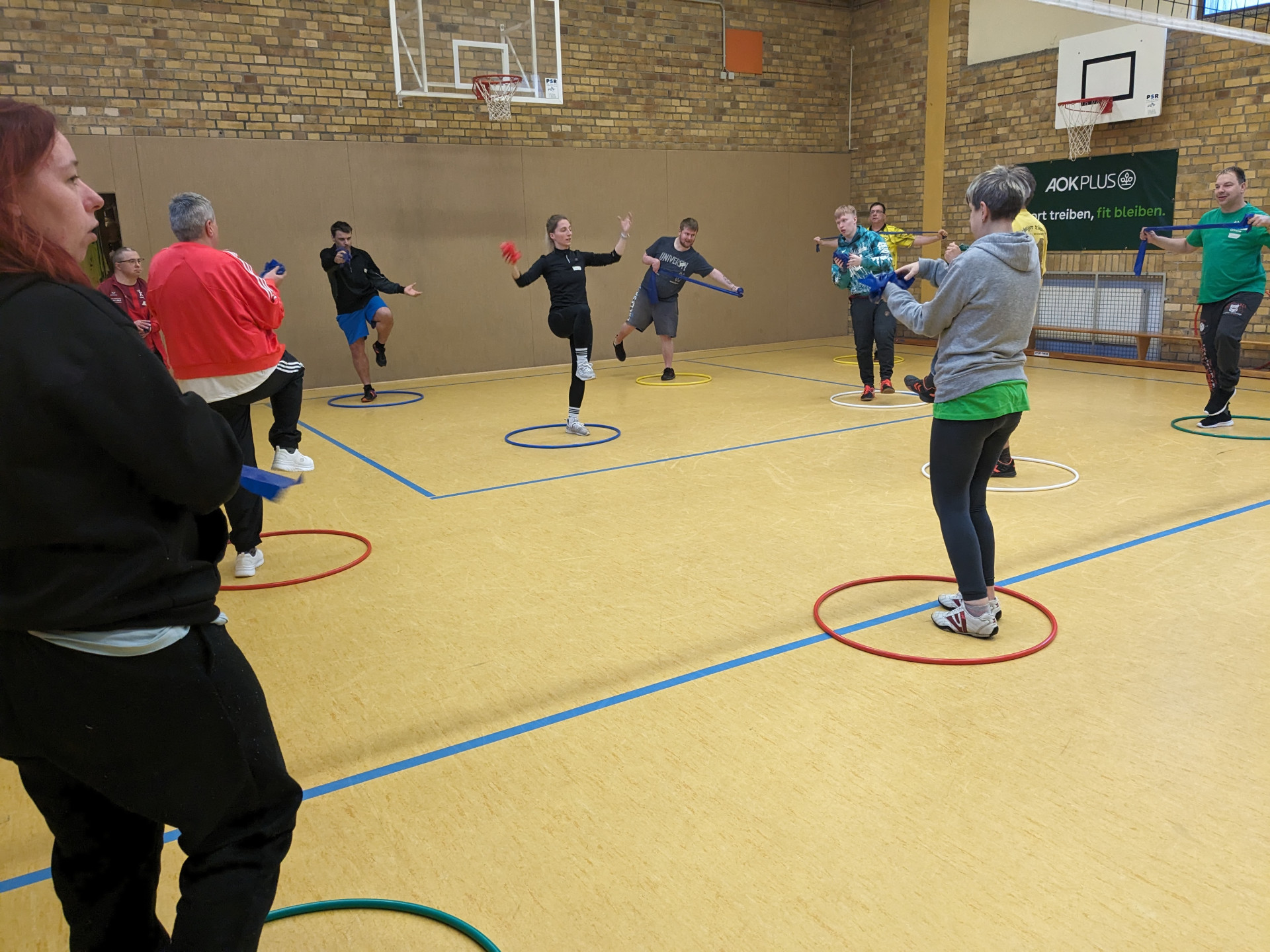 Es liegen Gymnastikreifen verteilt auf dem Hallenboden. Die Co-Trainer stehen in den Gymnastikreifen. Manche stehen auch auf einem Bein in den Gymnastikreifen.Sie halten ein Theraband in der Hand.