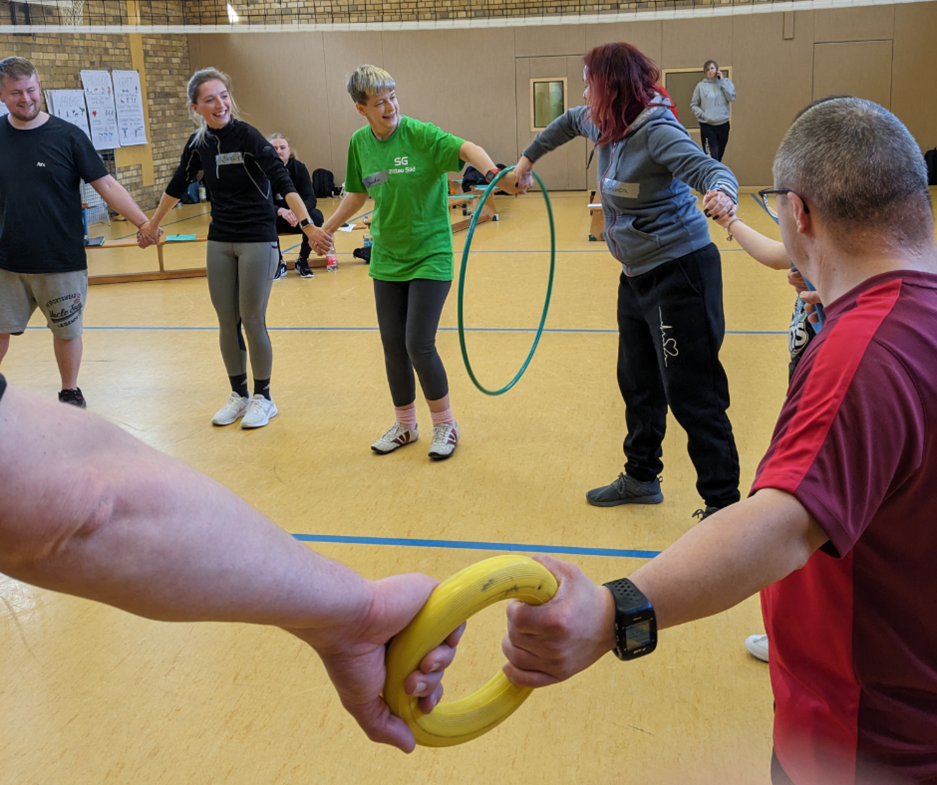 Die Co Trainer stehen in der Sporthalle und halten sich an den Händen. Sie bilden einen Kreis. Es ist ein Ausschnitt des Kreises zu sehen.  Alle haben Spaß.